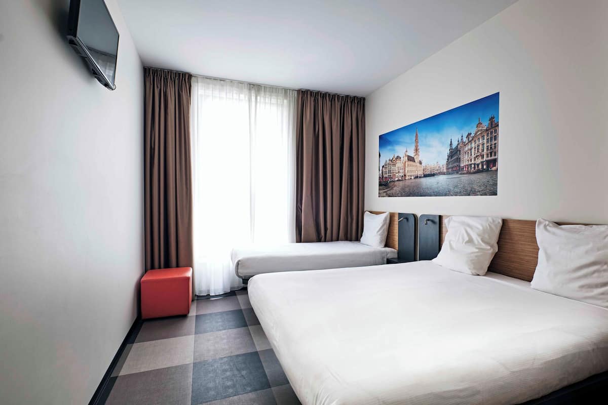 Ein Hotelzimmer mit Bett, einem an der Wand befestigten Fernseher, einem Fenster mit Vorhängen und einem Stadtbild.