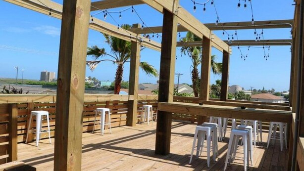 Terraza exterior con vigas de madera y taburetes blancos, con vistas a un estacionamiento y palmeras.