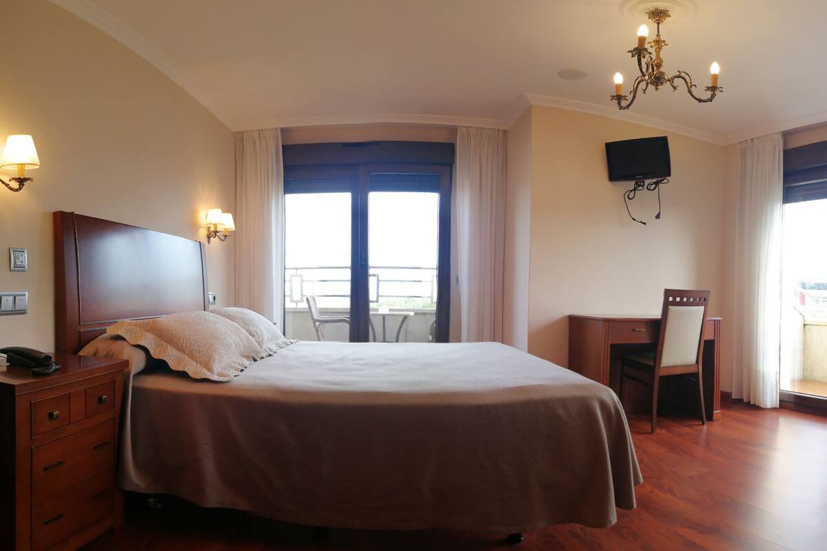 A hotel room with a large bed, a desk with a chair, a TV mounted on the wall, and a balcony with a view.
