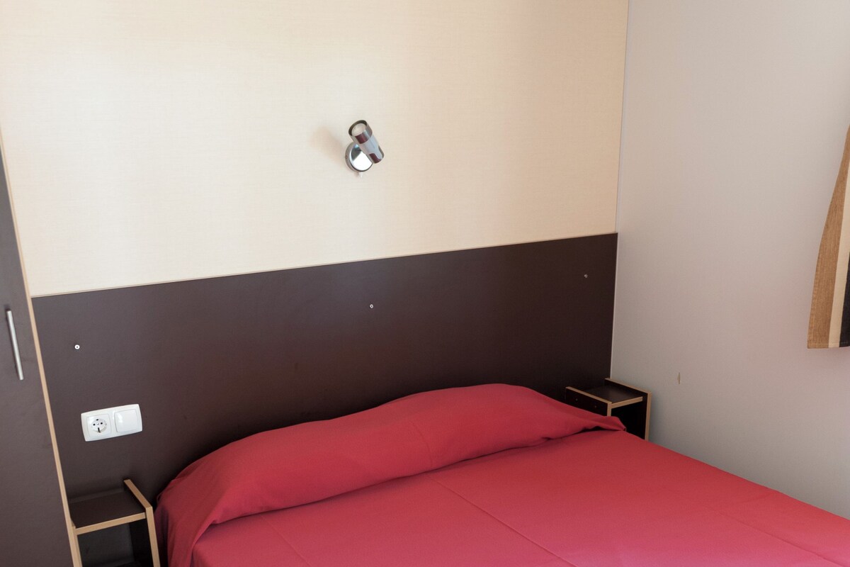 Ein Einzelbett mit roter Bettdecke in einem Zimmer mit braun-weißem Kopfteil, einem kleinen Holfnachtkästchen und einer wandmontierten Steckdose.
