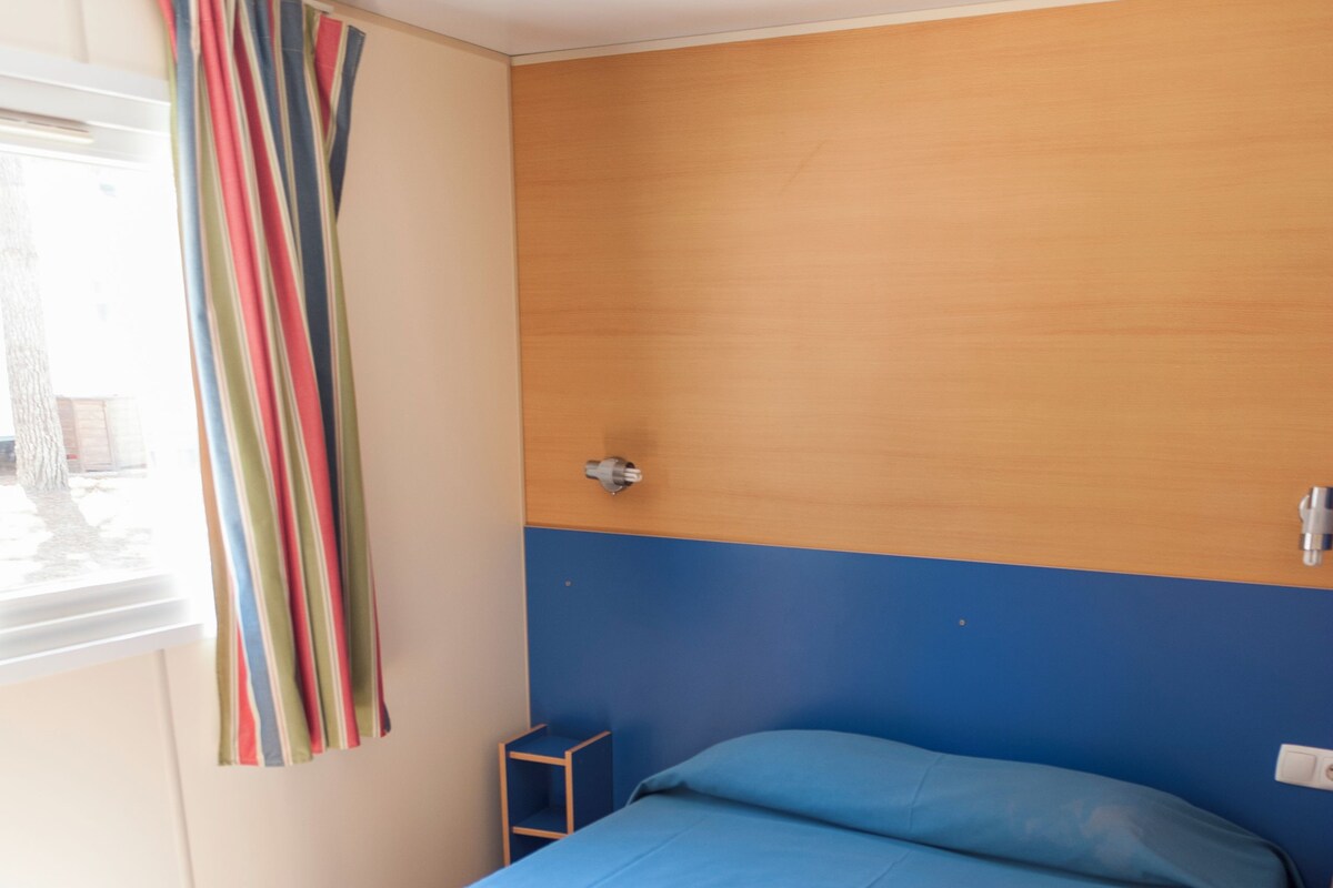 A single bed with a blue mattress in a room with wooden flooring and a window with a striped curtain.