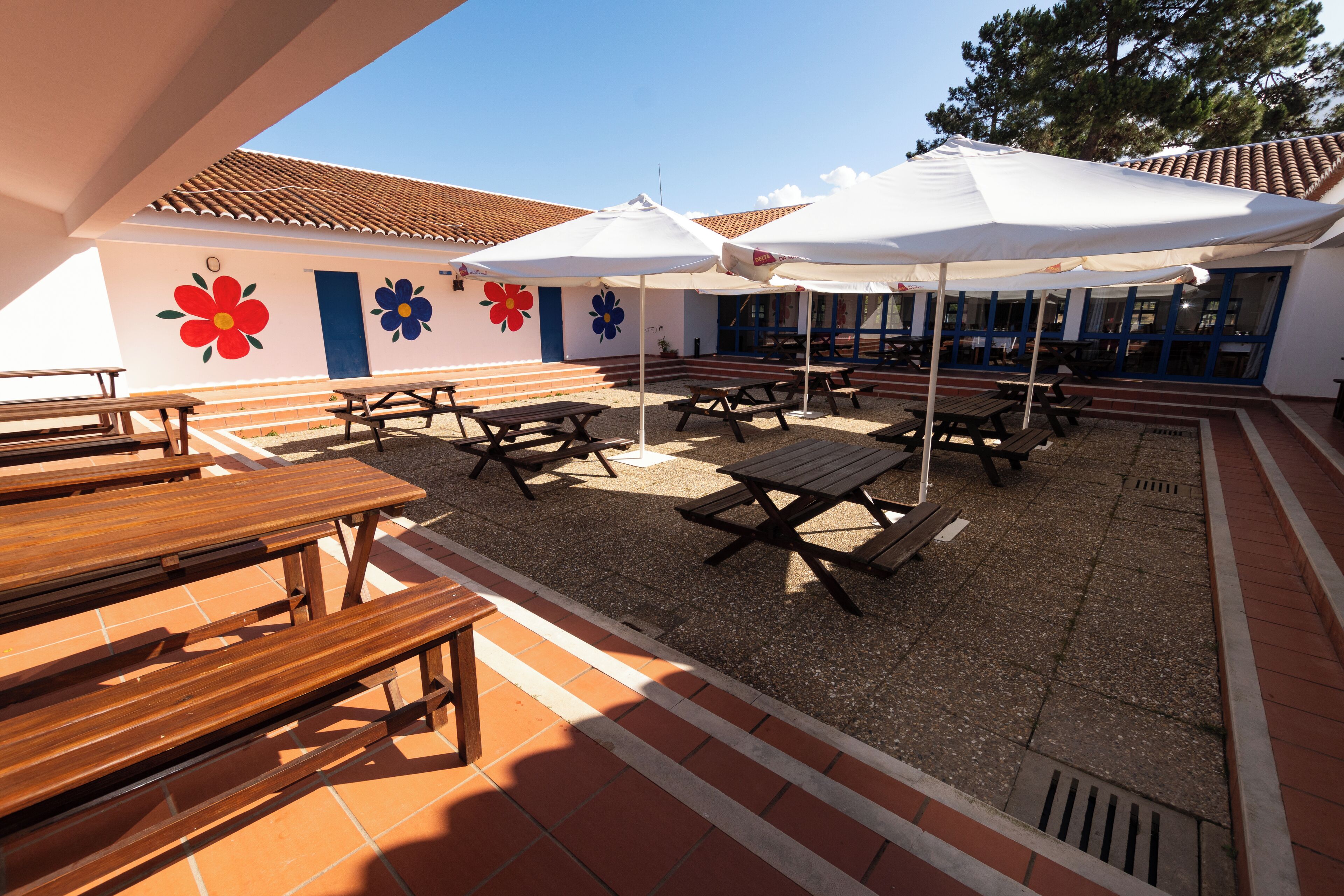 Ein Außenbereich mit einer Terrasse, Holztischen und -bänken, weißen Sonnenschirmen sowie einem Gebäude mit einem Ziegeldach und farbenfrohen Blumenmalereien.
