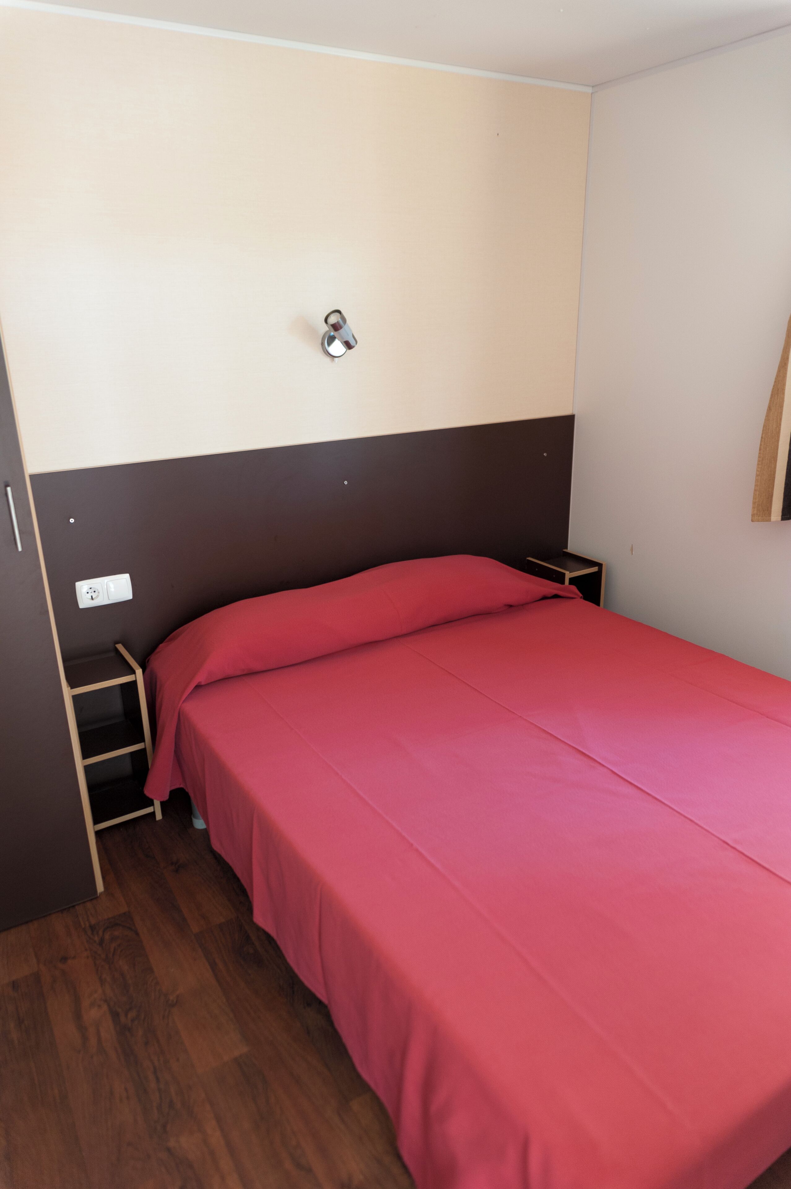 Ein Einzelbett mit roter Bettdecke in einem Zimmer mit braun-weißem Kopfteil, einem kleinen Holfnachtkästchen und einer wandmontierten Steckdose.