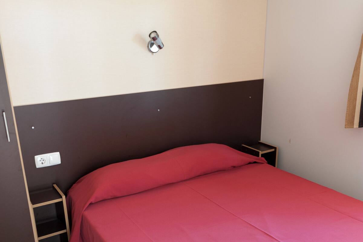 Ein Einzelbett mit roter Bettdecke in einem Zimmer mit braun-weißem Kopfteil, einem kleinen Holfnachtkästchen und einer wandmontierten Steckdose.