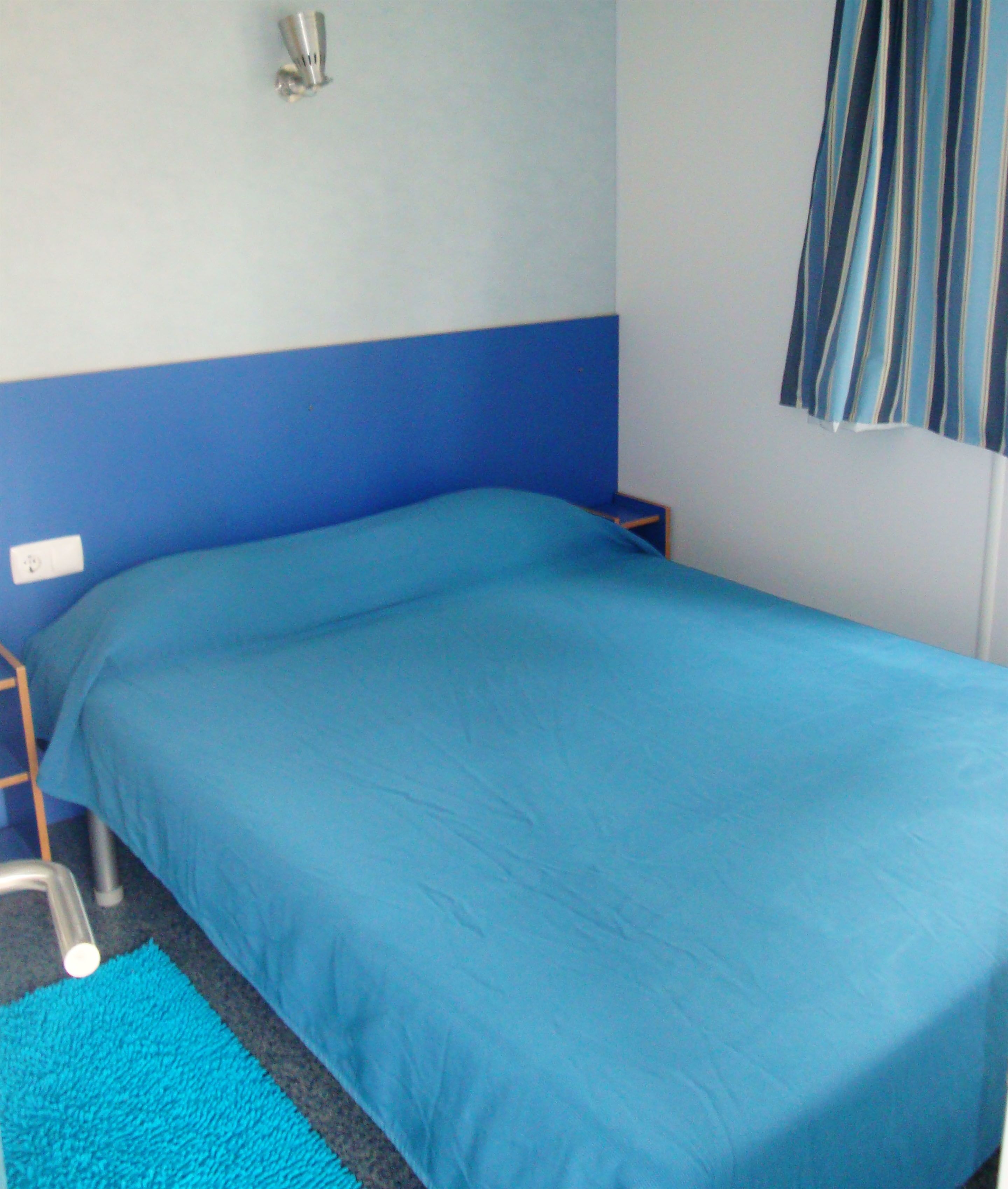 Une chambre avec un lit simple, un mur bleu, une fenêtre avec des rideaux bleus et une petite table de chevet.