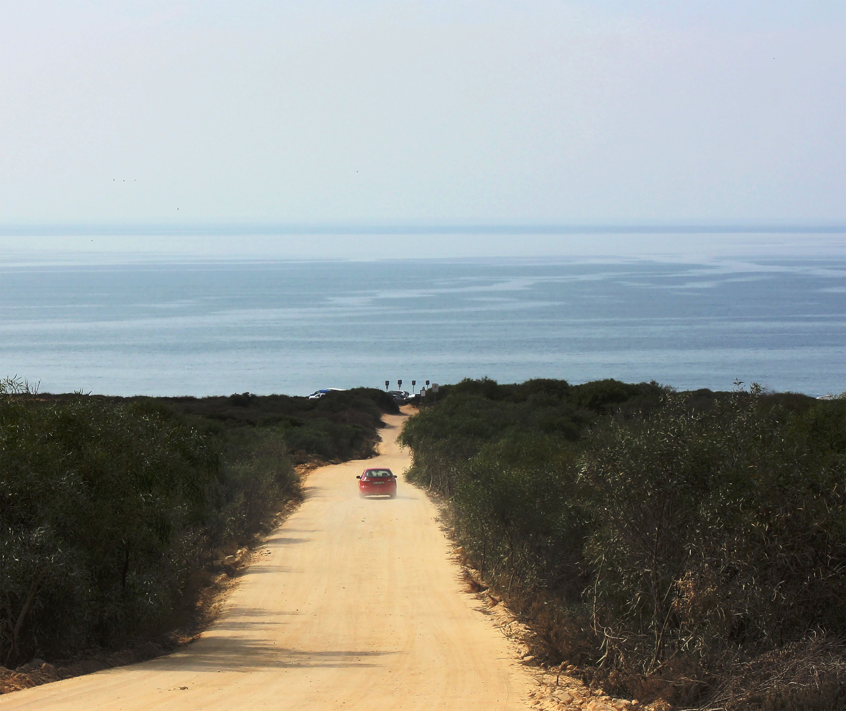 Un camino de tierra que lleva a una playa, con un coche rojo estacionado al borde.