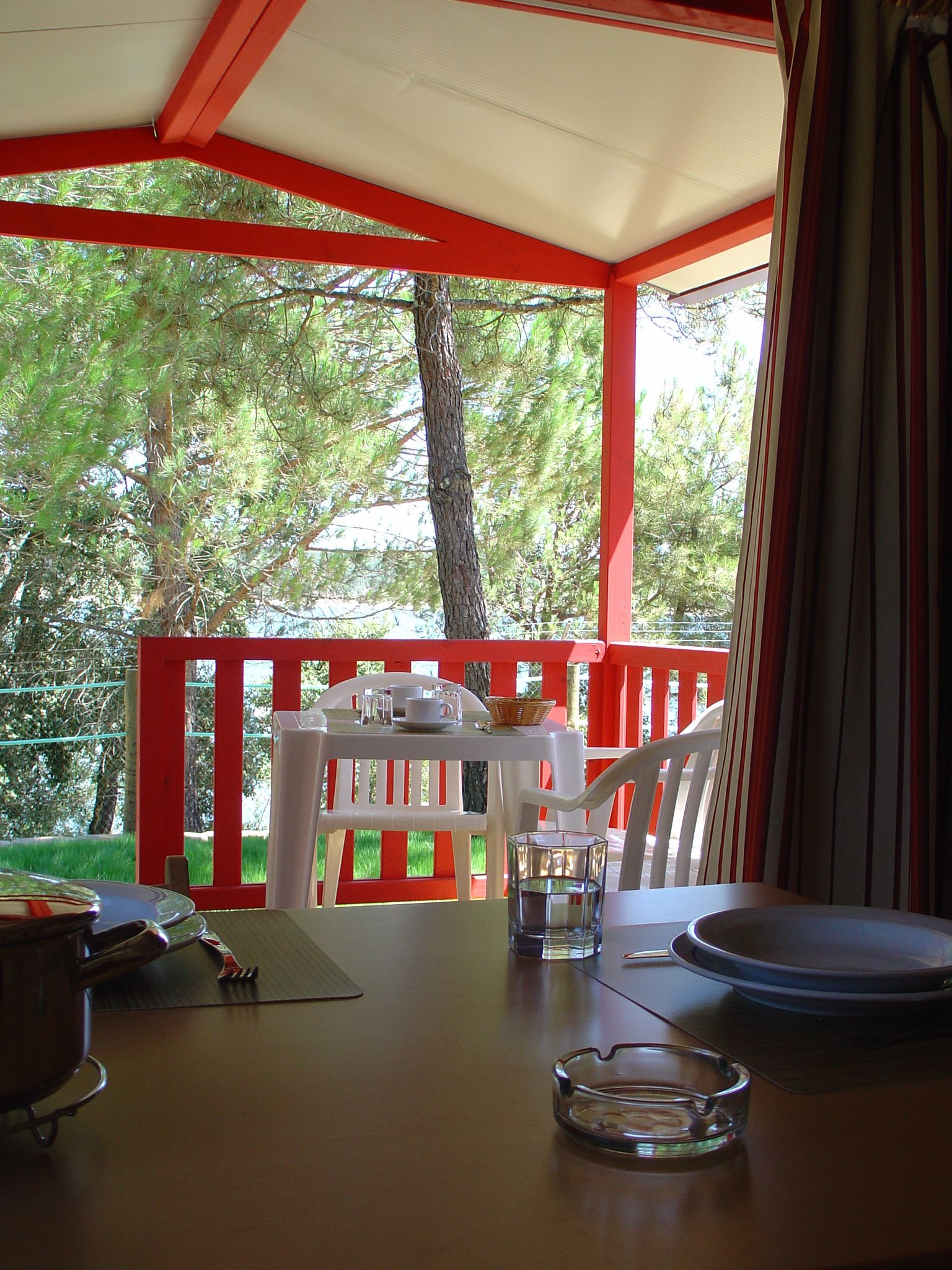 A covered outdoor seating area with a red railing, a table with chairs, and a view of trees.