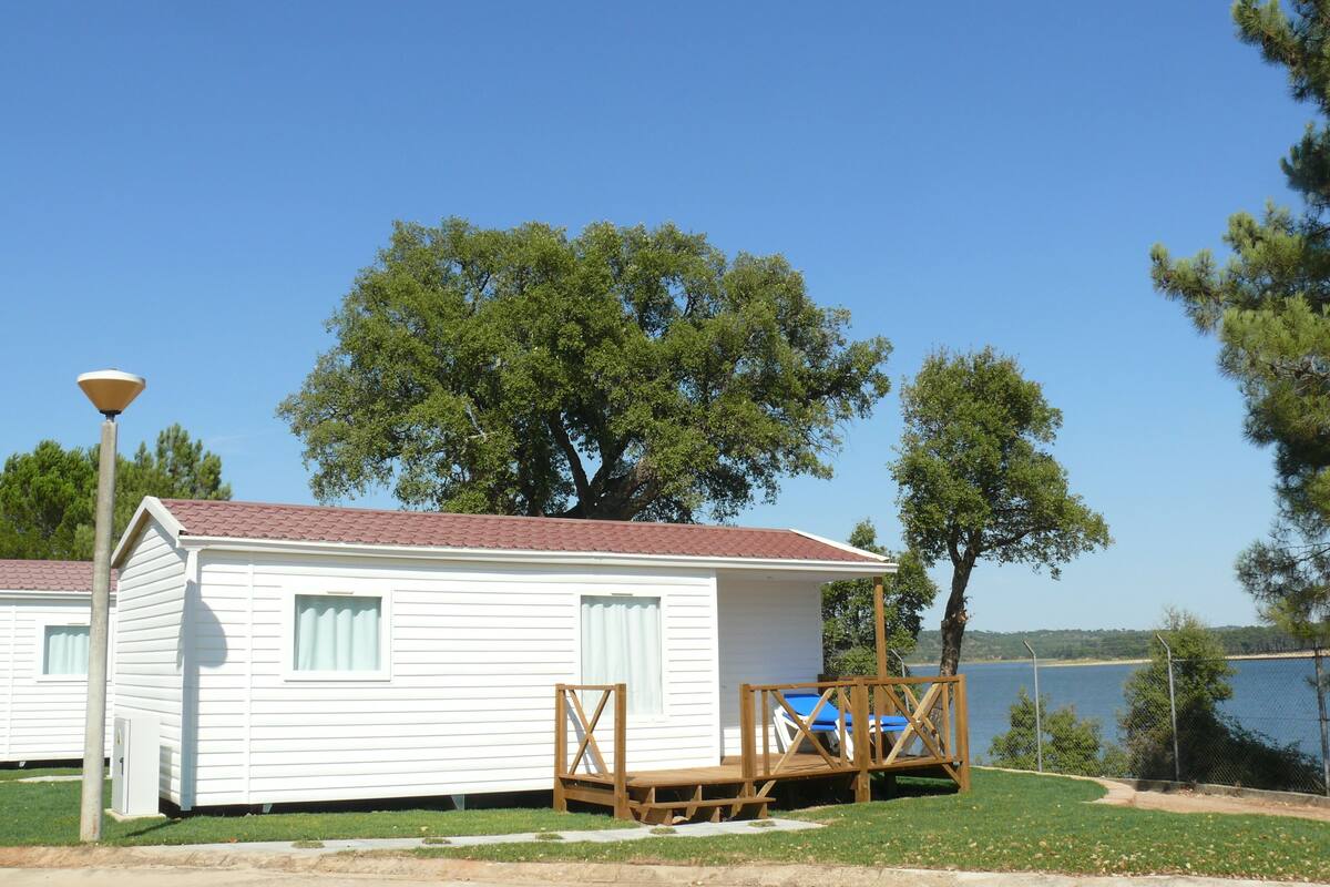 A small white cabin with a red roof, a wooden deck, and a view of a lake.