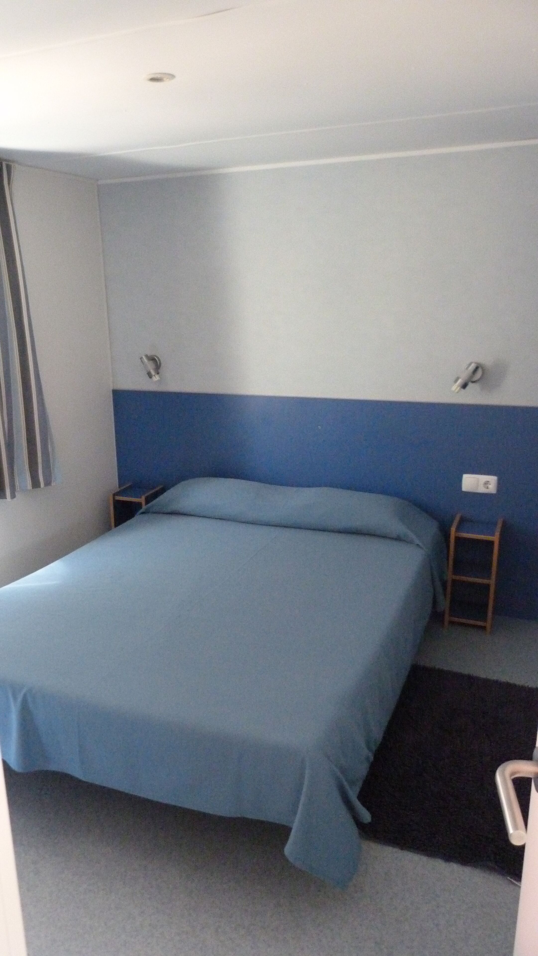 A single bed with a blue bedspread in a room with light blue and white walls, a small wooden nightstand, and a black rug on the floor.