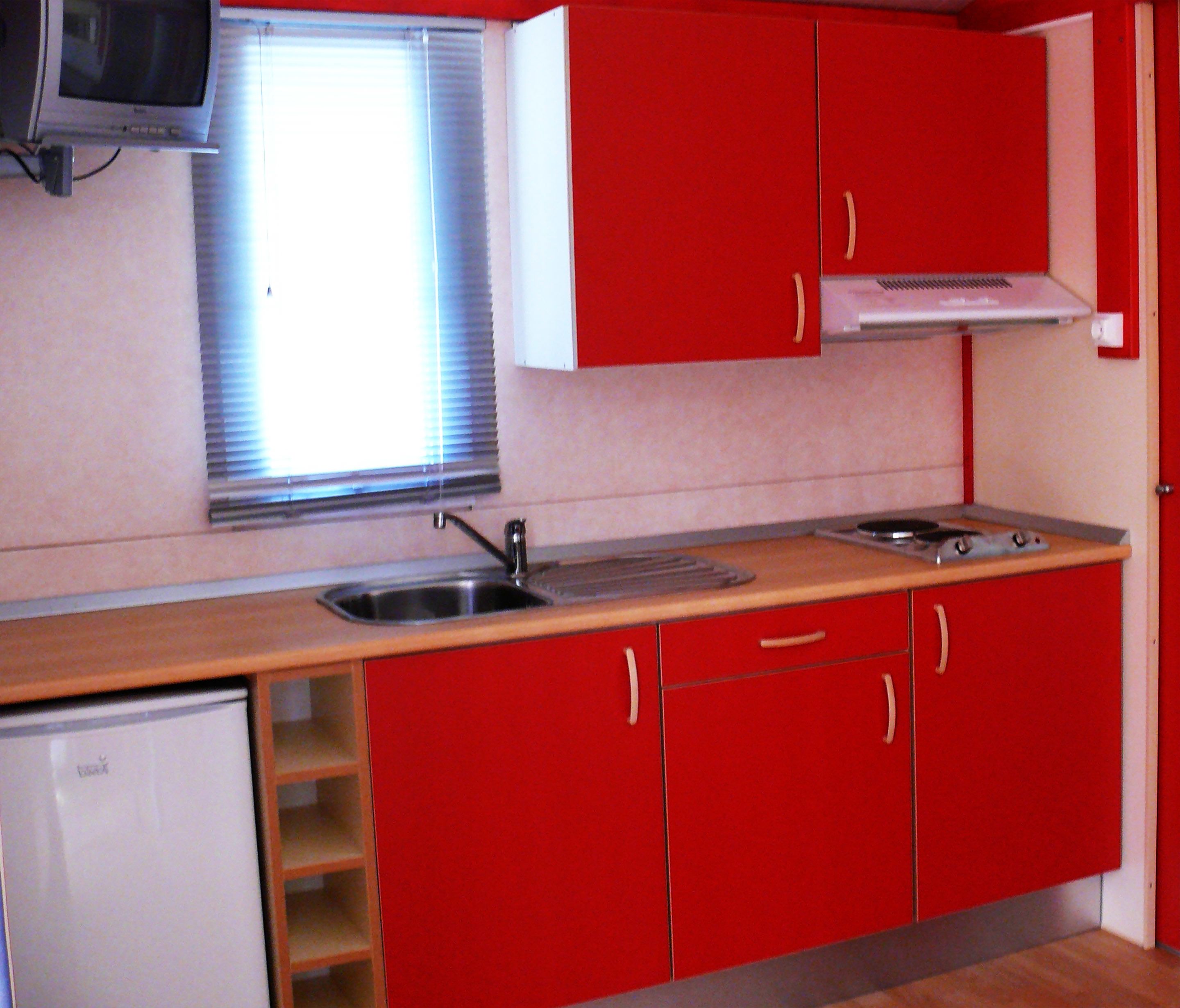 A compact kitchen with red cabinets, a sink, a small refrigerator, and a window with blinds.
