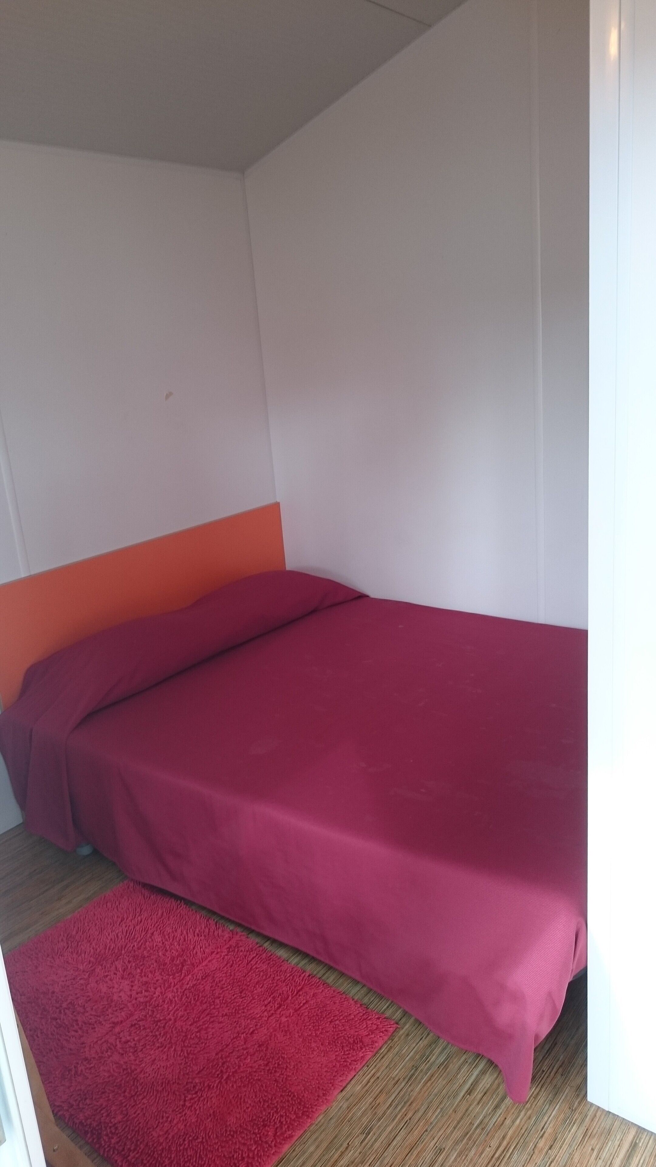A single bed with a red mattress and orange headboard in a room with white walls and a wooden floor.