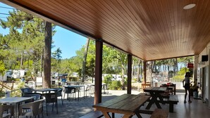 An outdoor patio area with wooden tables and chairs, a covered seating area, and a view of the surrounding trees and parked vehicles. - Parque de Campismo Orbitur Caminha (Caminha)
