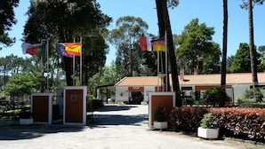 A building entrance with multiple flags, a paved area, and a fence with greenery. - Parque de Campismo Orbitur Caminha (Caminha)