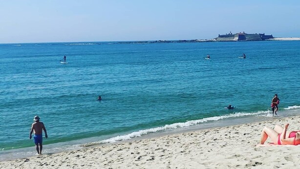 A beach with clear blue water, a sandy shore, and a distant fort-like structure. - Parque de Campismo Orbitur Caminha (Caminha)