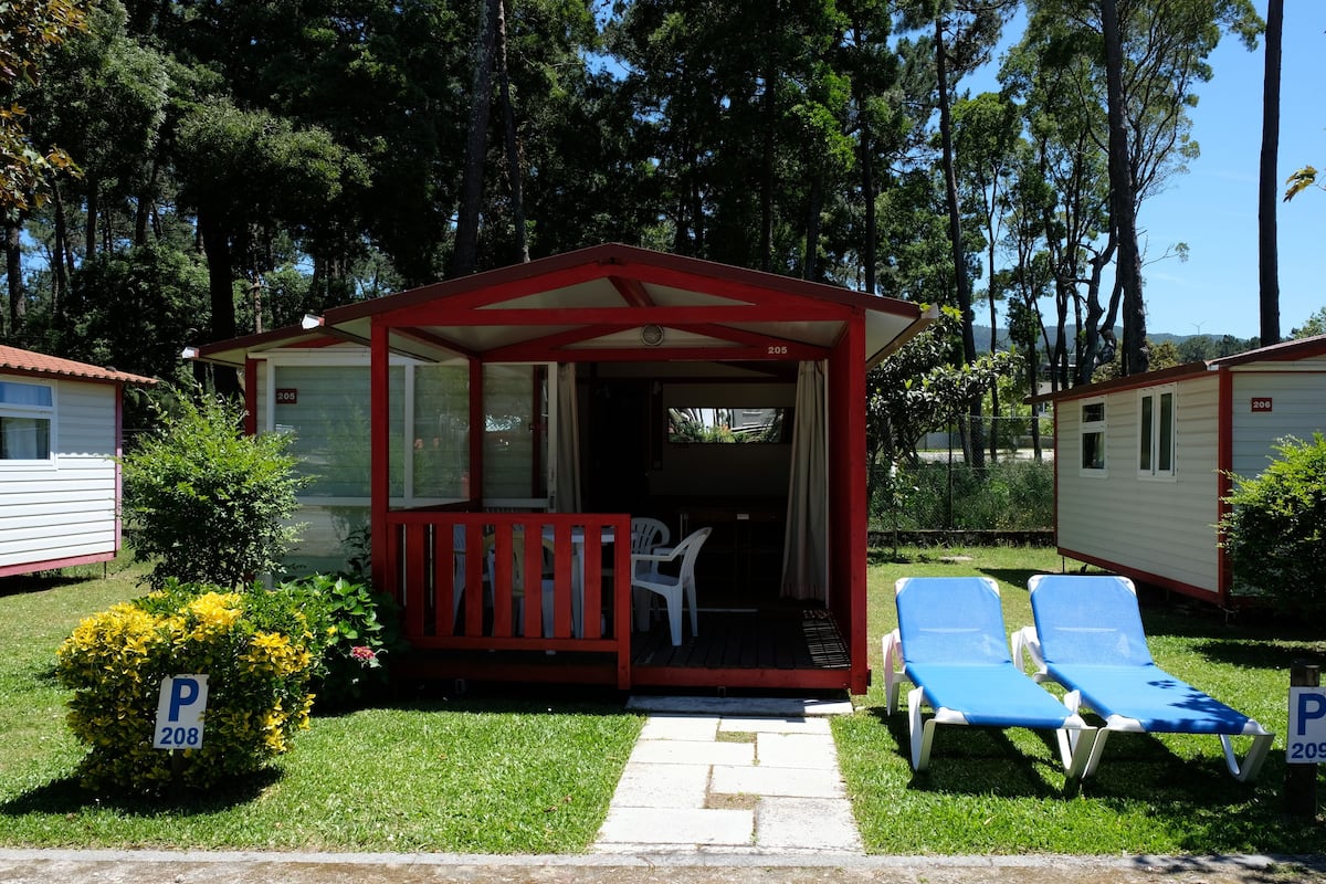 A small, red and white mobile home with a porch, two blue lounge chairs, and a small garden area.