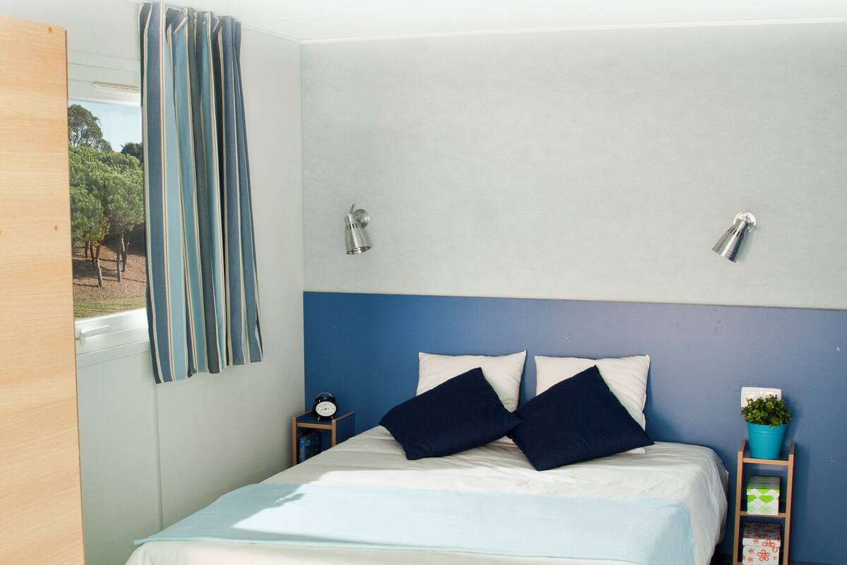 Une chambre à coucher avec un lit, une table de chevet, une lampe fixée au mur, une fenêtre avec des rideaux et une petite plante posée sur une table d’appoint.