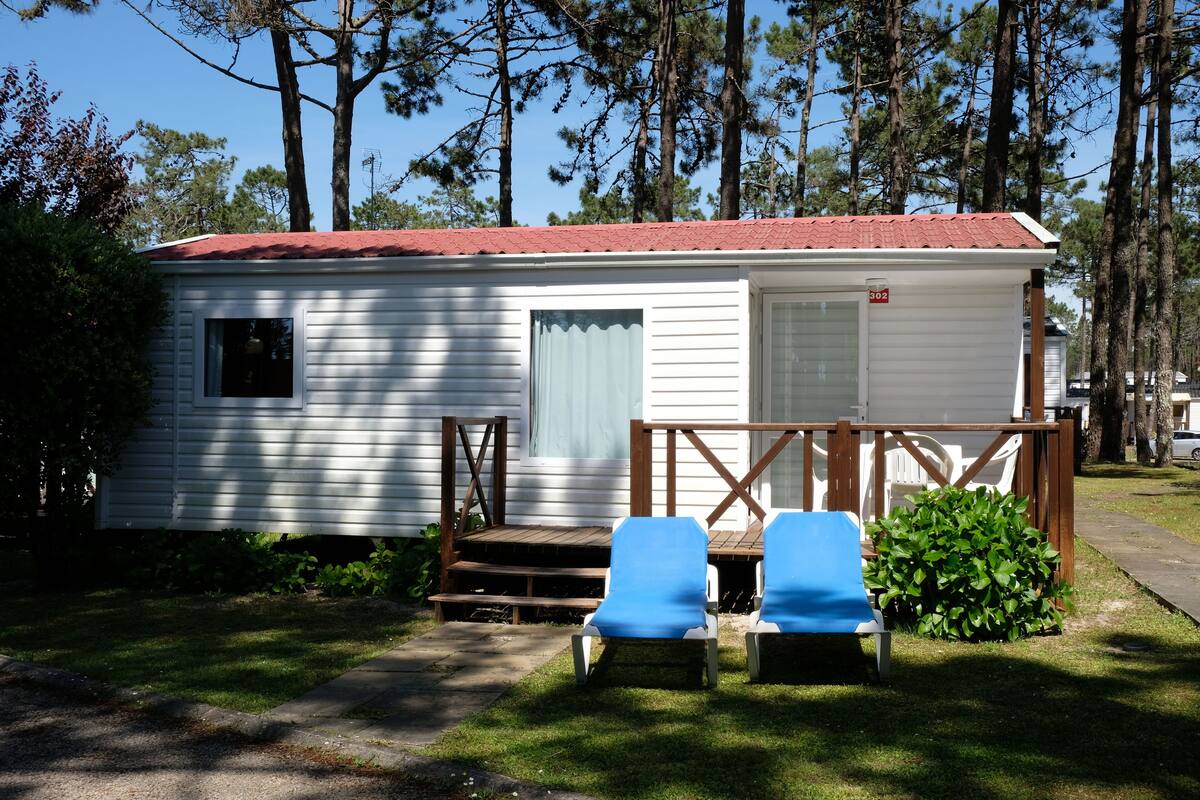 Une petite maison mobile blanche avec un toit rouge, deux chaises de jardin bleues et une terrasse en bois.