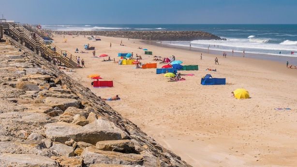 Ein Strand mit felsiger Küste, einer Holztreppe und bunten Strandsonnenschirmen.