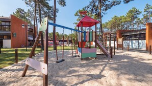 Ein Spielplatz mit Schaukeln und einer Kletterkonstruktion, umgeben von Sand und Bäumen.