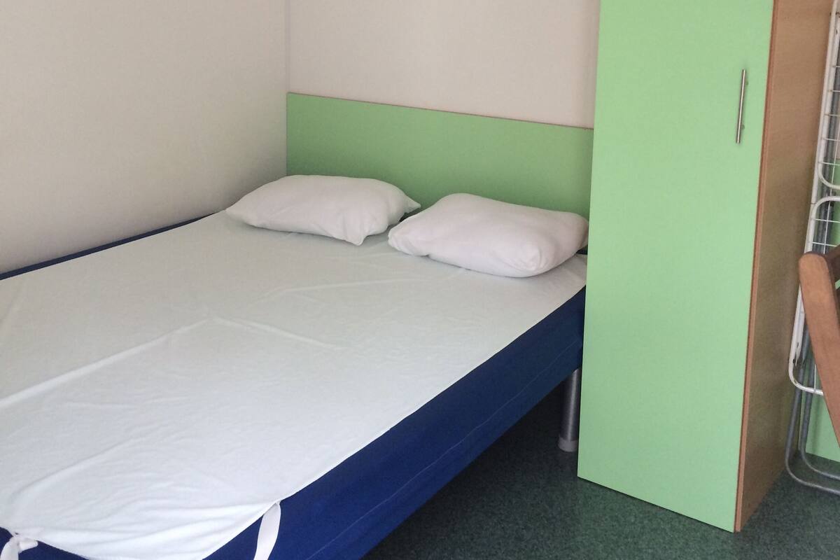 Un lit simple avec un cadre bleu et une literie blanche, une tête de lit verte et une armoire verte dotée d’une poignée en métal.