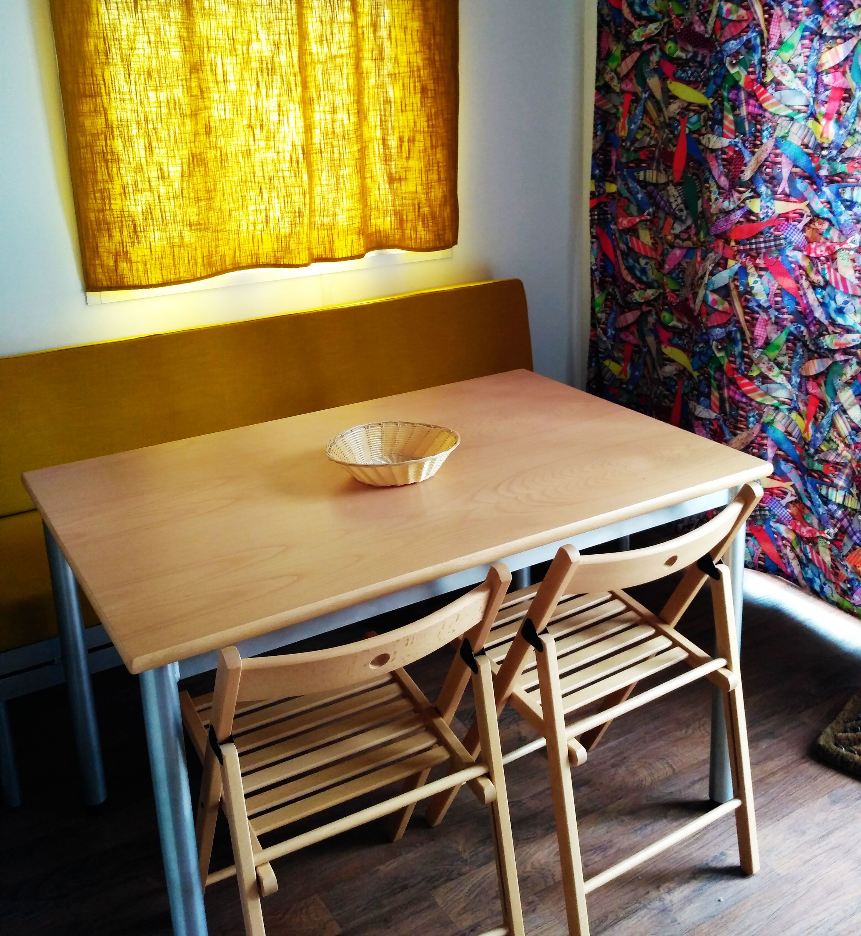 A small wooden table with a woven basket, two wooden chairs, a bench, and a colorful curtain.