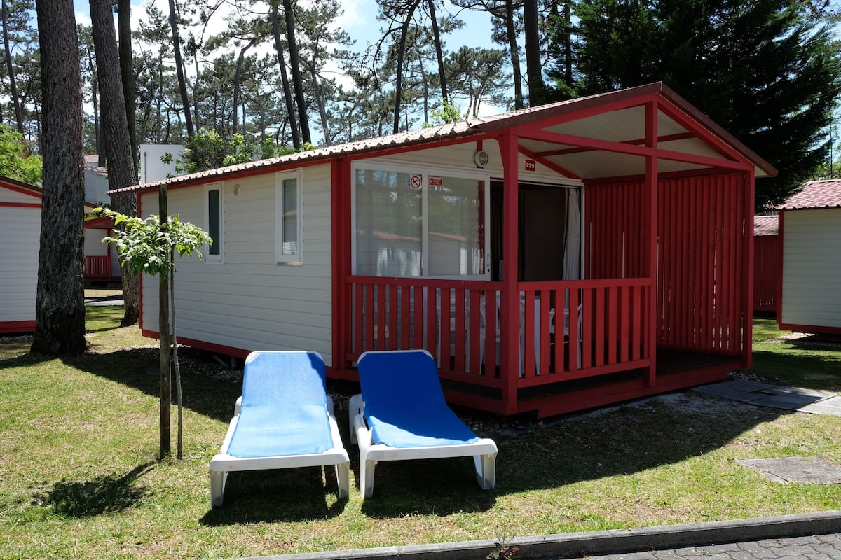 A small, single-story cabin with a red exterior and a covered porch. There are two blue lounge chairs outside.