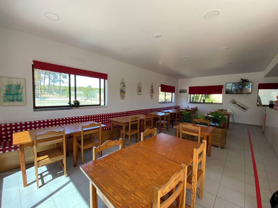 Restaurante com mesas e bancos de madeira, grandes janelas com cortinas vermelhas e uma televisão na parede.