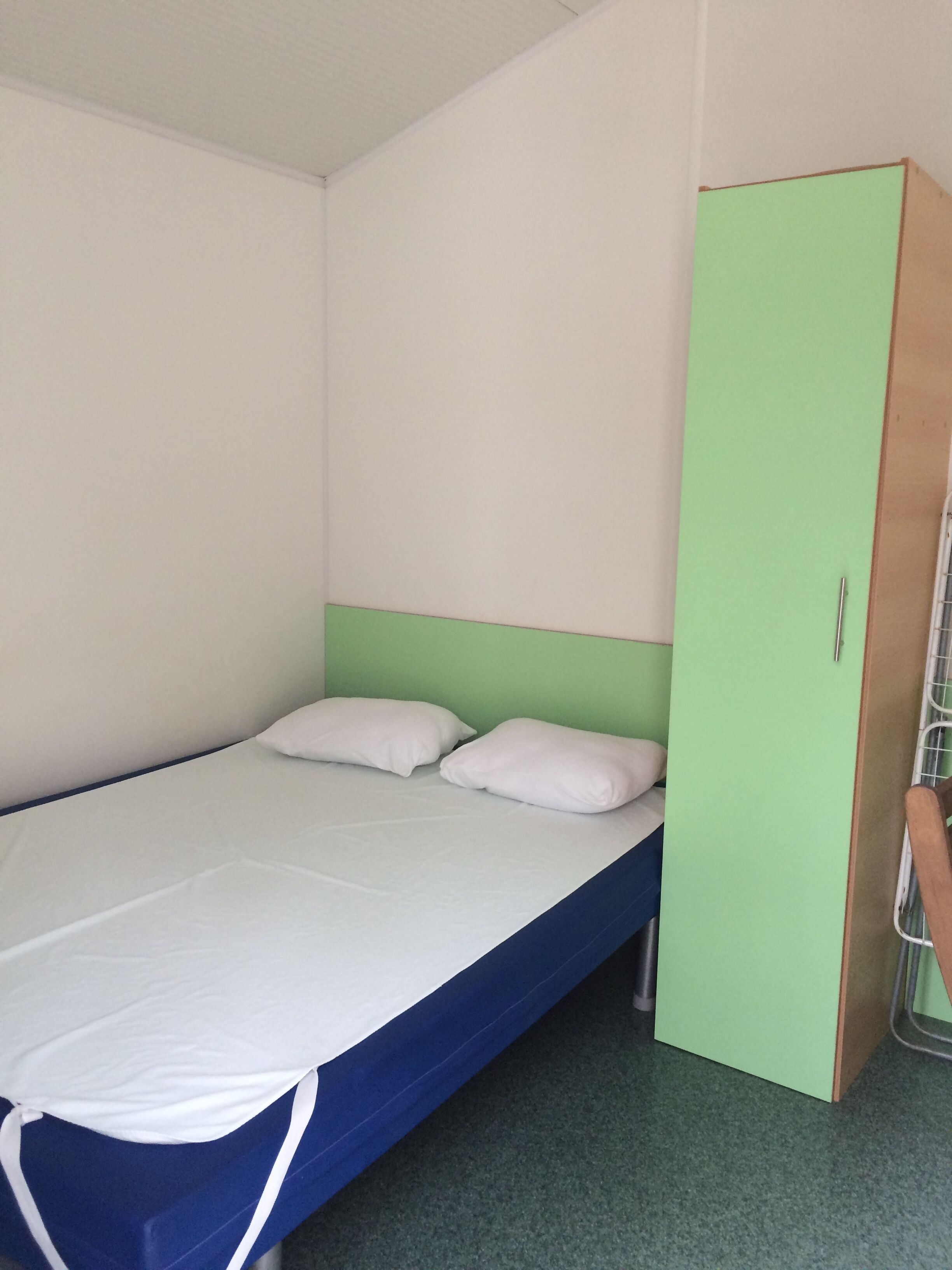 Una cama individual con estructura azul, ropa de cama blanca, cabecera verde y armario verde con pomo metálico.