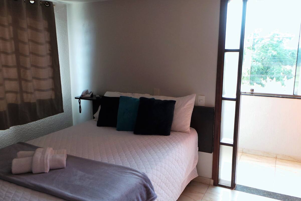 A bedroom with a bed, pillows, a bedside table with a phone, and a window with a view of greenery.