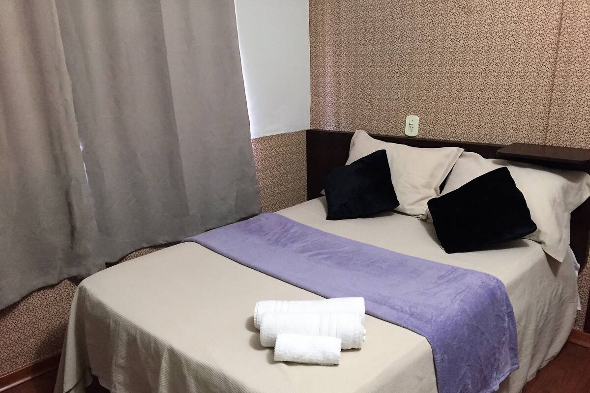 A neatly made bed with a beige bedspread, a purple throw blanket, and two pillows (one black and one white). There are two white towels folded on the bed. The room has a carpeted floor and a window with curtains.