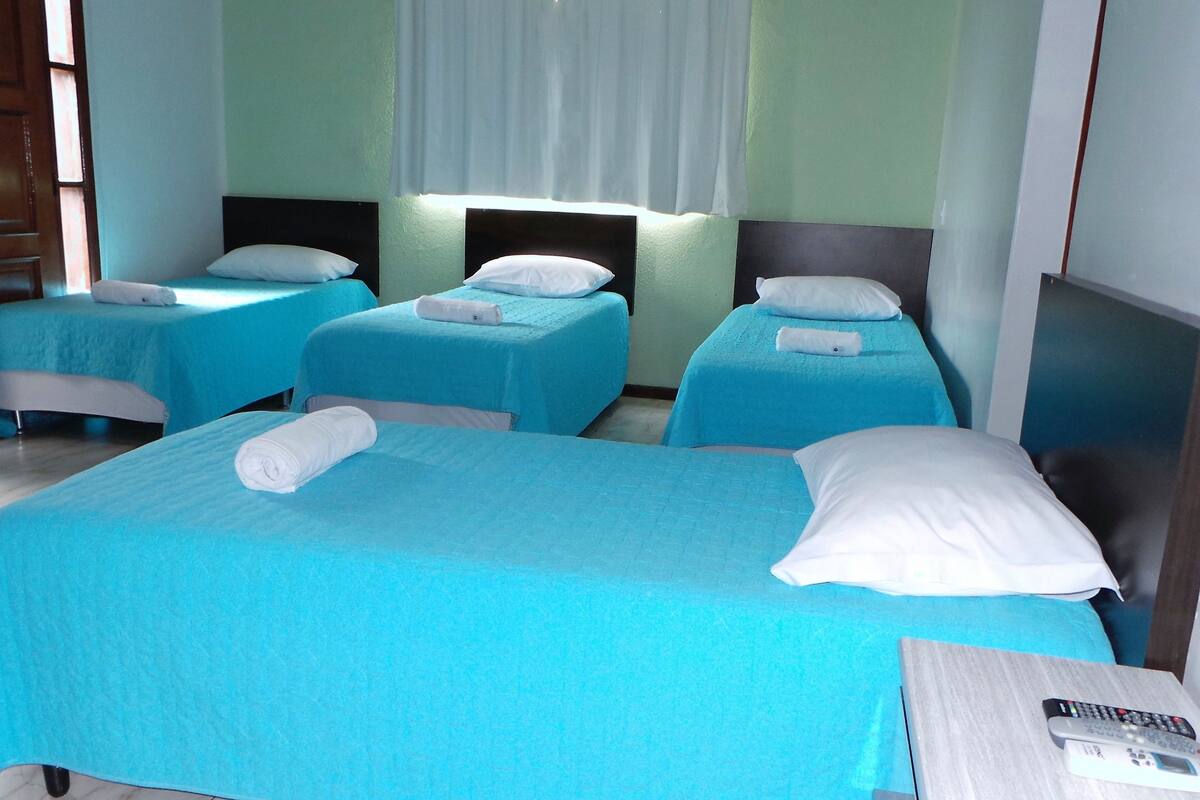A hotel room with two single beds and one double bed, each with blue bedding and white pillows. There is a wooden door on the left and a mirror on the right wall.