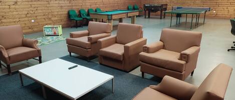A room with wooden walls, a pool table, green chairs, and a ping pong table.