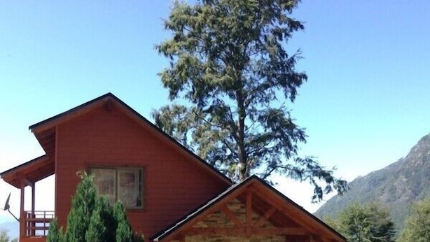 A red wooden cabin with a stone porch, surrounded by greenery and mountains. - Cabaña Fau Wualmapu (Pucón)