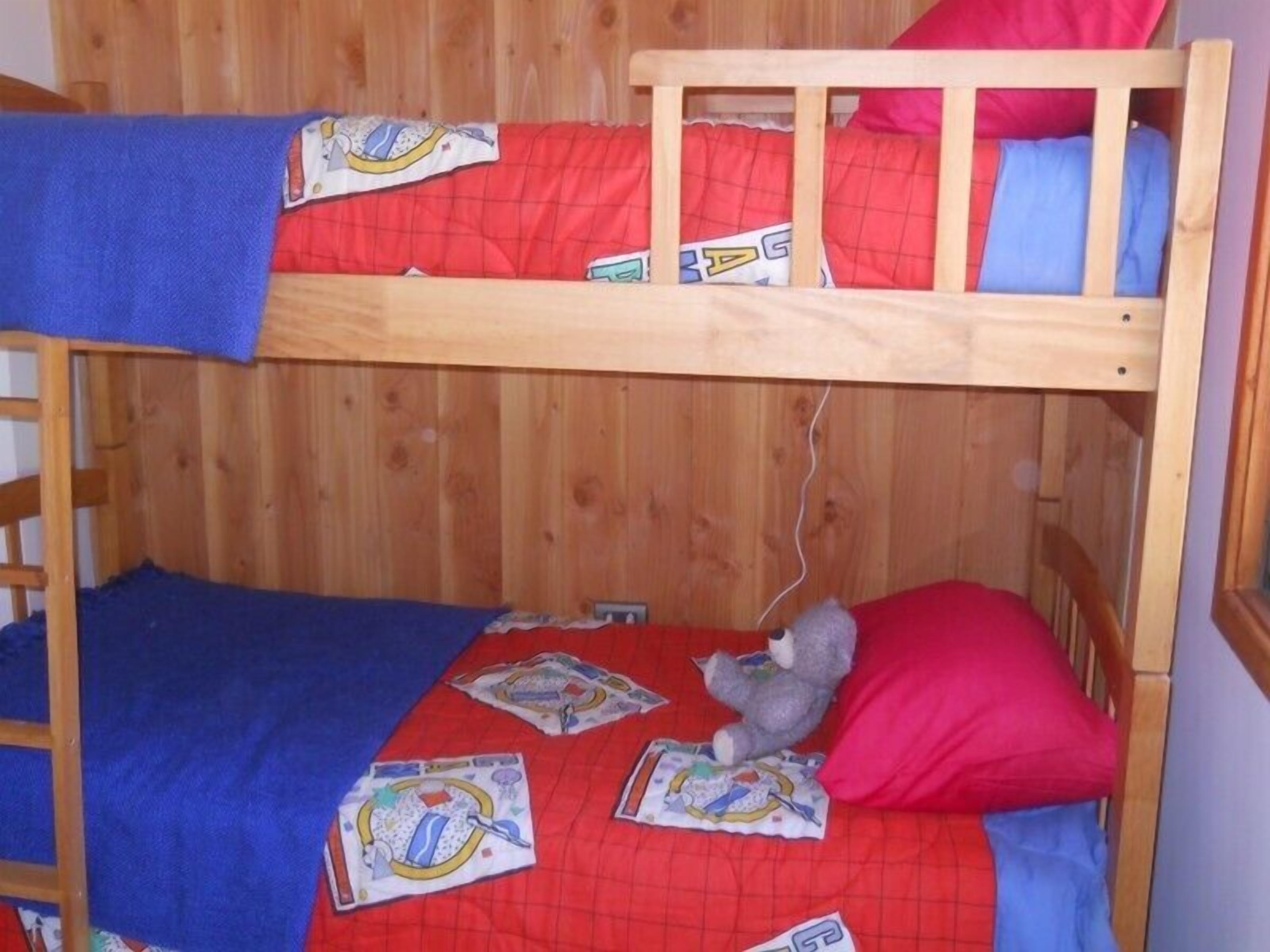 Ein Etagenbett mit Holzrahmen, roter und blauer Bettwäsche und einem Stofftier auf der unteren Ebene.