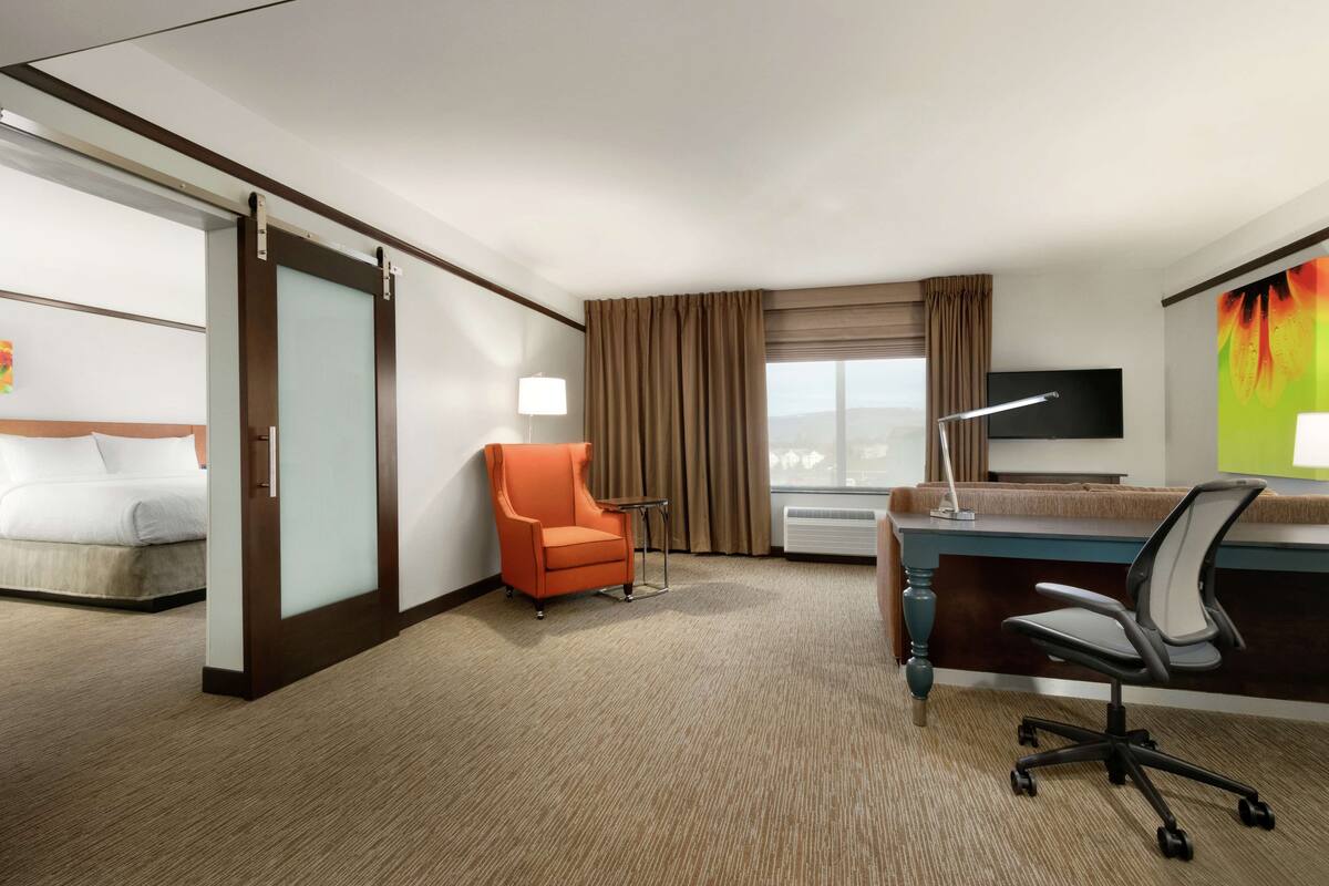 Ein Hotelzimmer mit Bett, orangefarbenem Sessel, Schreibtisch mit Stuhl, Flachbildfernseher und einem Fenster mit Vorhängen.