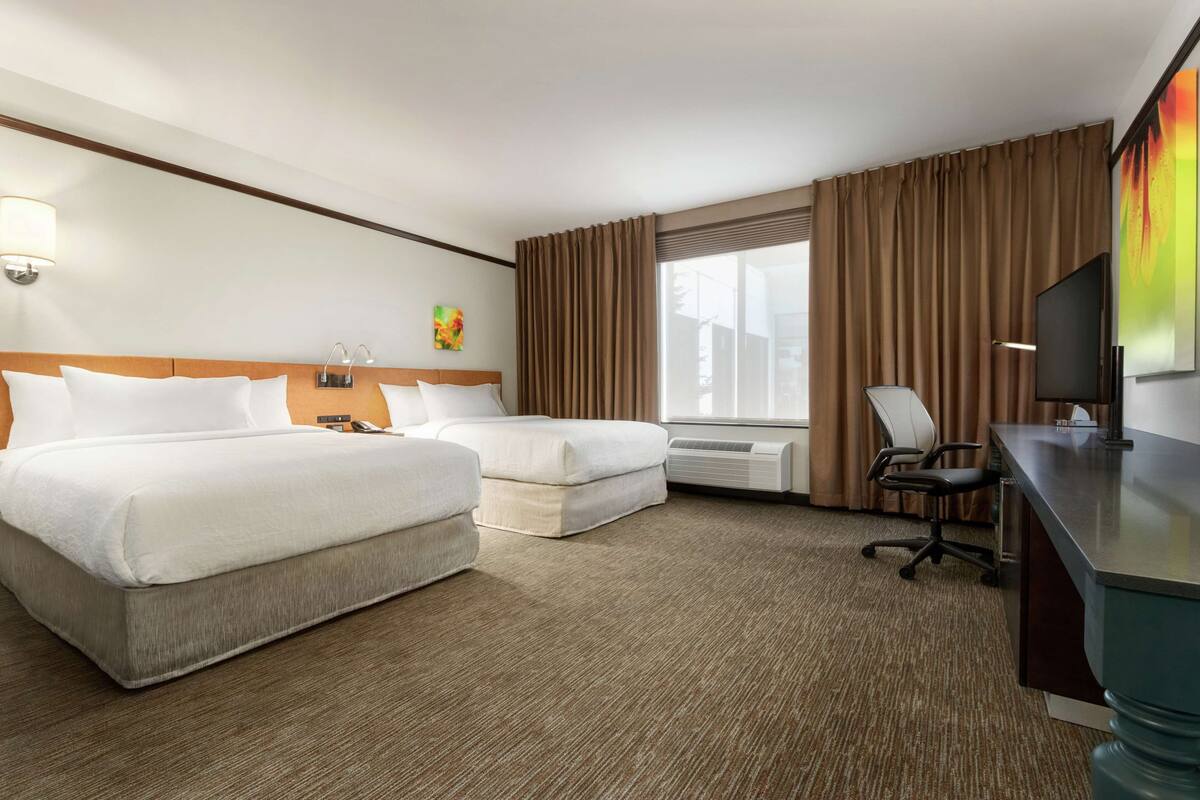 Ein Hotelzimmer mit zwei Betten, einem Schreibtisch, einem Stuhl, einem Fernseher und einem Fenster mit Vorhängen.