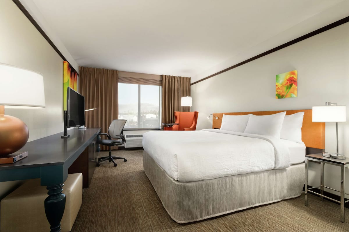 Ein Hotelzimmer mit einem großen Bett, einem Schreibtisch mit Computer, einem Stuhl und einem Fenster mit Vorhängen.
