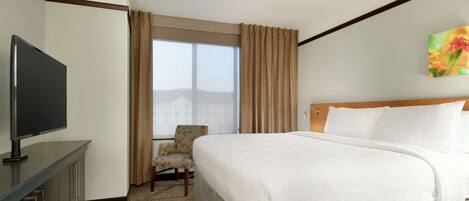 A hotel room with a bed, a TV, a chair, and a window with curtains.