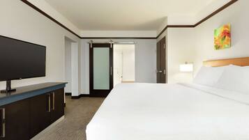 Una habitación de hotel con una cama grande, un televisor de pantalla plana sobre un mueble oscuro, una lámpara de noche y una puerta que da a otra habitación.