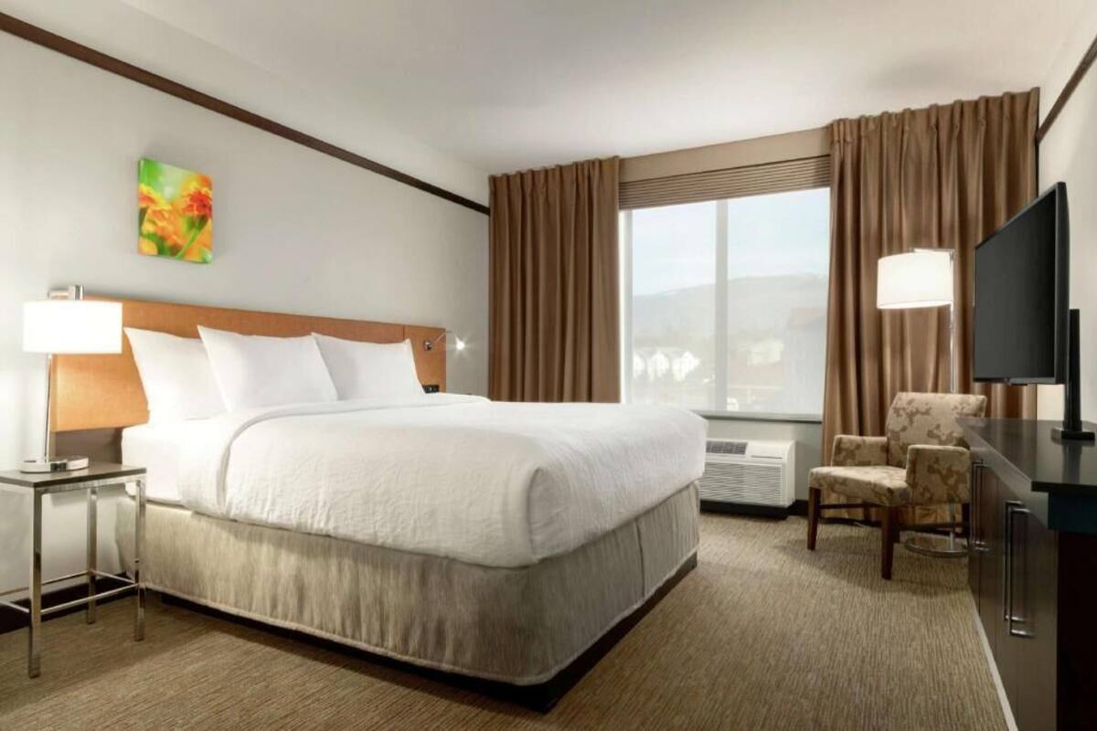 Ein Hotelzimmer mit einem großen Bett, einem Schreibtisch mit Lampe, einem Fernseher, einem Sessel und einem Fenster mit Vorhängen.