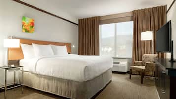 A hotel room with a large bed, a desk with a lamp, a chair, a television, and a window with curtains.