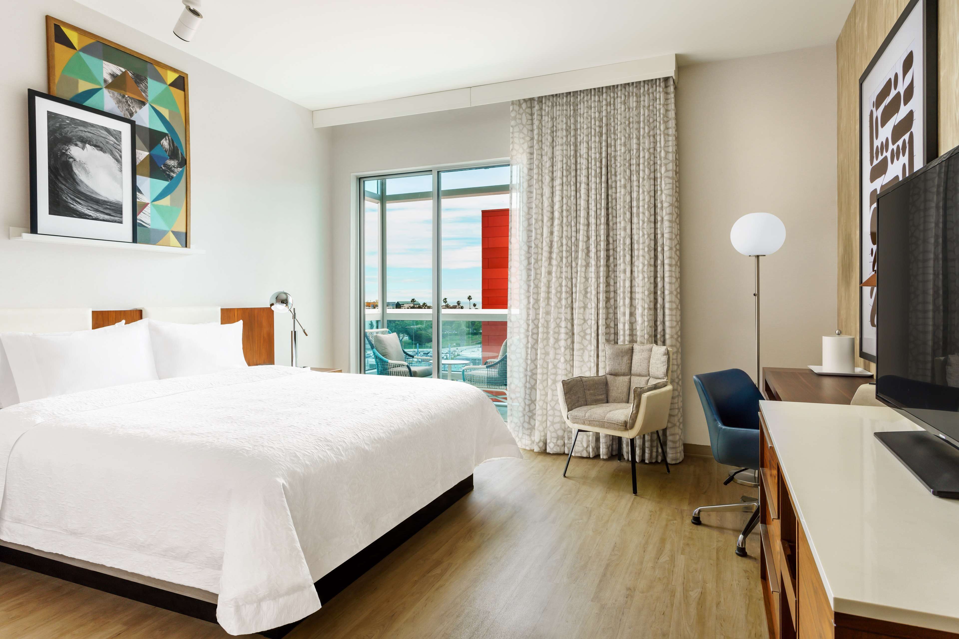 A hotel room with a large bed, a desk with a TV, a chair, and a view of the outdoors through a large window.