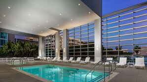 Une piscine intérieure à l’eau d’un bleu cristallin, entourée de chaises longues blanches et dotée d’un mur de verre offrant une vue sur le ciel nocturne.