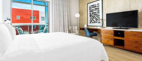 A modern hotel room with a large bed, a flat-screen TV, a wooden cabinet, and a view of a red building through a large window.