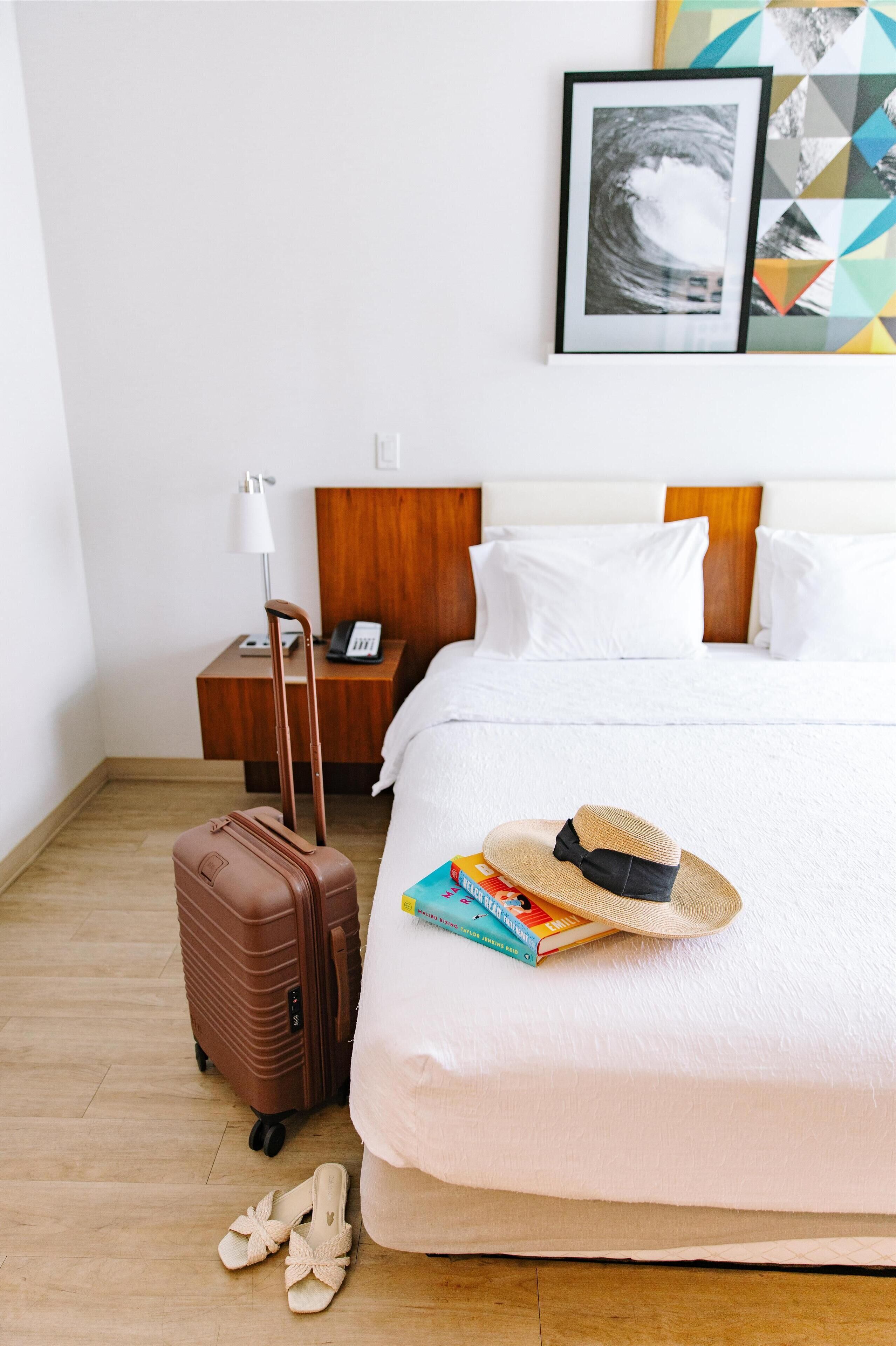 Une chambre d’hôtel avec un lit, une table de chevet avec une lampe, une valise, une paire de chaussons et un chapeau posé sur le lit.