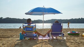 Two beach chairs under a blue umbrella, a beach ball, and a person walking in the background. - Barren River Lake State Resort Park (Lucas)