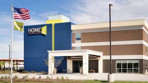 A modern hotel building with a prominent "HOME 2 Suites by Hilton" sign, a covered entrance, and a flagpole with the American flag.