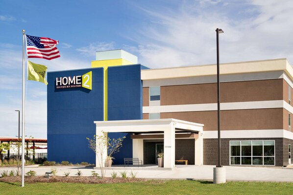 A modern hotel building with a prominent "HOME 2 Suites by Hilton" sign, a covered entrance, and a flagpole with the American flag.
