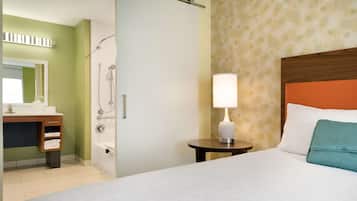 Habitación de hotel con cama, mesita de noche con lámpara, baño con ducha y tocador con espejo.
