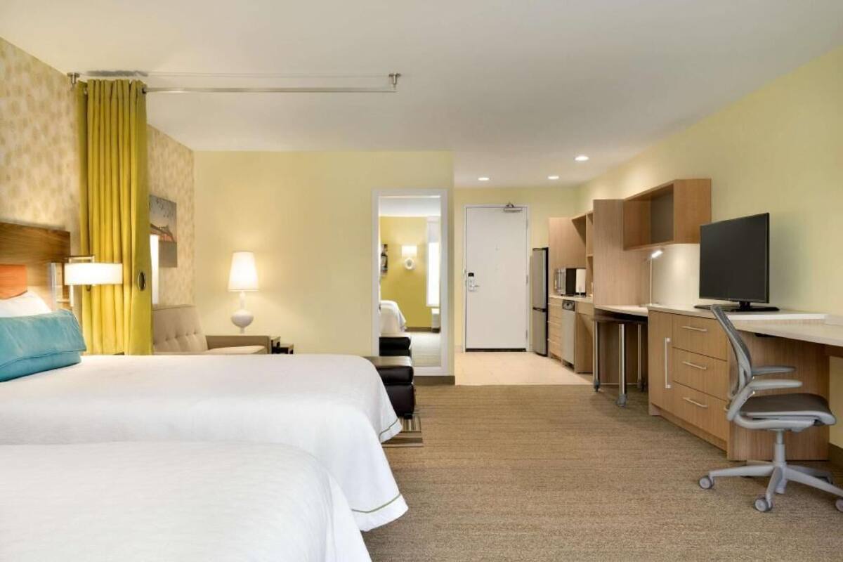 A hotel room with two beds, a desk, a television, and a kitchenette.